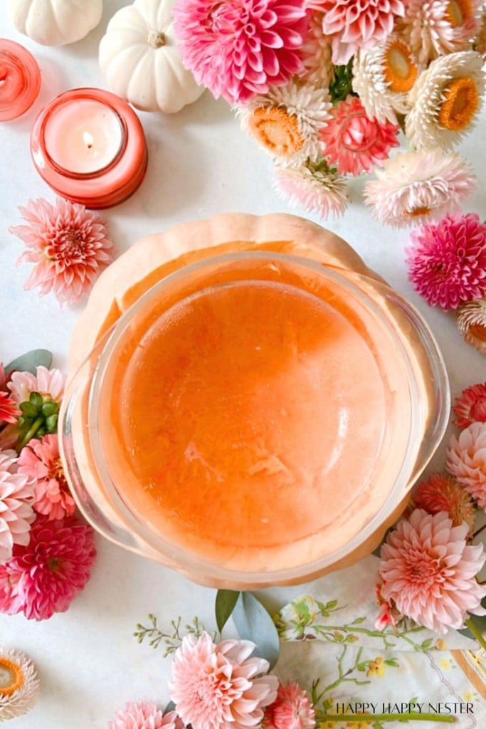 A round transparent bowl filled with orange liquid sits on a table surrounded by pink and white flowers and small candles. The background includes a mix of colorful flowers and small white pumpkins, creating a vibrant and cozy atmosphere.