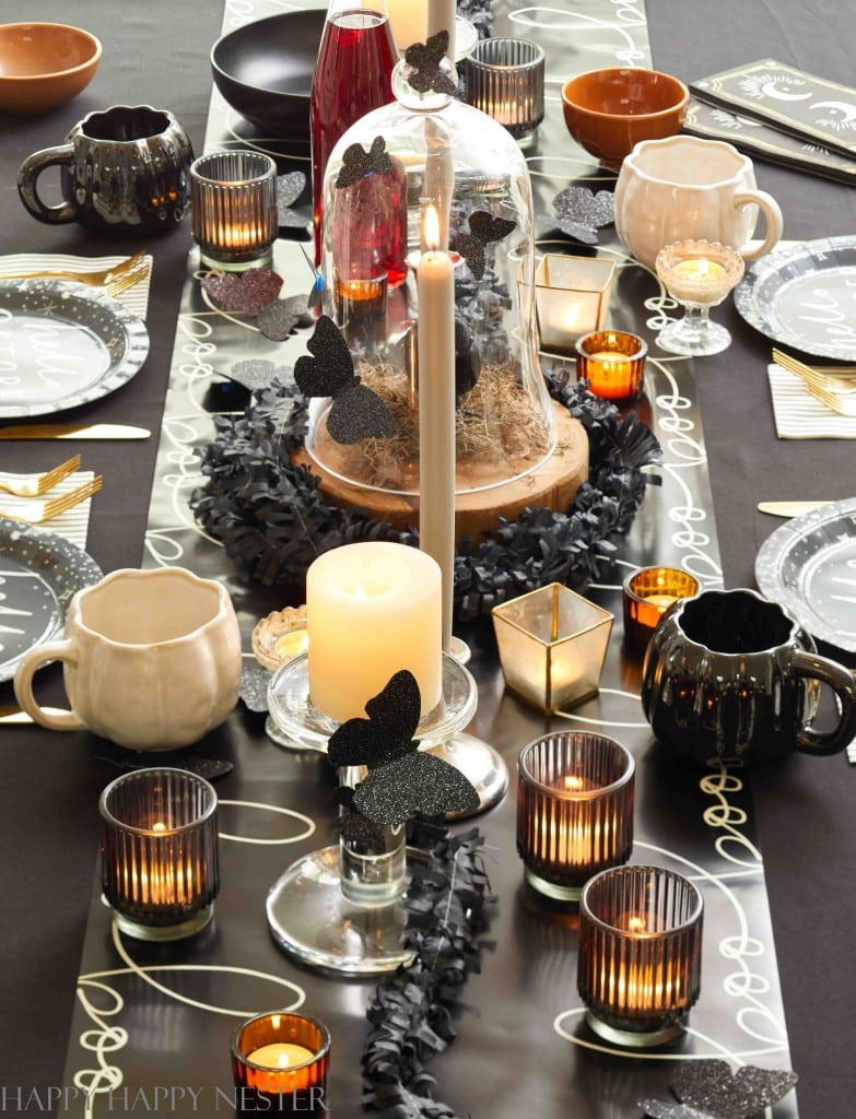 A stylish Halloween-themed table setting features a black and gold table runner with a candle centerpiece adorned with black butterflies. Various candleholders, mugs shaped like cauldrons, orange and white cups, and decorative bats accent the scene.