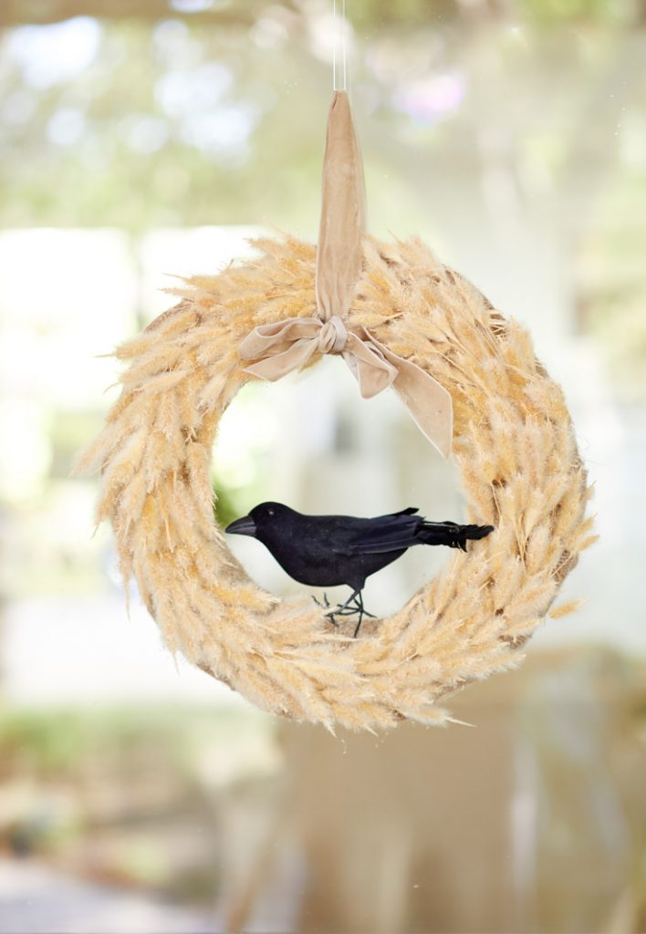 A decorative wreath made of beige, feathery materials is hanging against a blurred background. A plush black crow is perched inside the wreath, adding a touch of contrast to the soft, neutral tones. The festive fall decor is suspended by a beige ribbon tied in a bow.