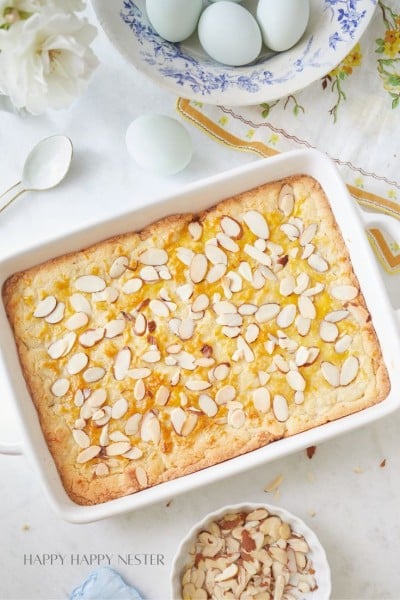 A baked dessert topped with almond slices in a rectangular white dish. Next to it are a spoon, a small bowl filled with almonds, and a plate of light blue eggs. The scene is set on a white surface with a floral-patterned cloth partially visible.