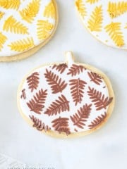 A decorated cookie shaped like a pumpkin is covered in white icing and adorned with brown fern-like leaf patterns. The cookie sits on a light surface alongside other round cookies with similar yellow leaf patterns. Text at the bottom reads "HAPPY HAPPY NESTER.