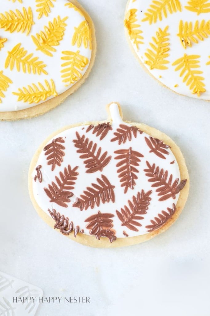 A decorated cookie shaped like a pumpkin is covered in white icing and adorned with brown fern-like leaf patterns. The cookie sits on a light surface alongside other round cookies with similar yellow leaf patterns. Text at the bottom reads "HAPPY HAPPY NESTER.