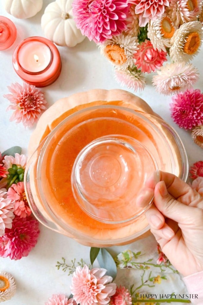 A top-down view of a hand holding a glass bowl over a larger pink fluid-filled bowl, surrounded by vibrant pink, white, and orange flowers. Various candles and decorative pumpkins are scattered around, creating a cozy and festive ambiance.