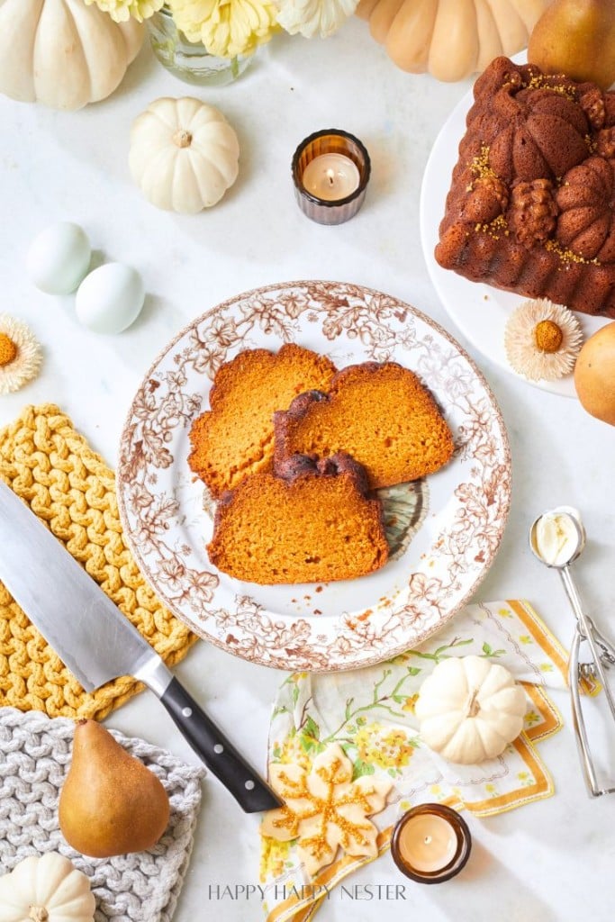 A fall-themed table setting featuring slices of pumpkin bread on a decorative plate, surrounded by autumnal items including white and yellow pumpkins, a knife, pears, candles, and knitted pot holders. A larger portion of pumpkin bread is in the back corner.