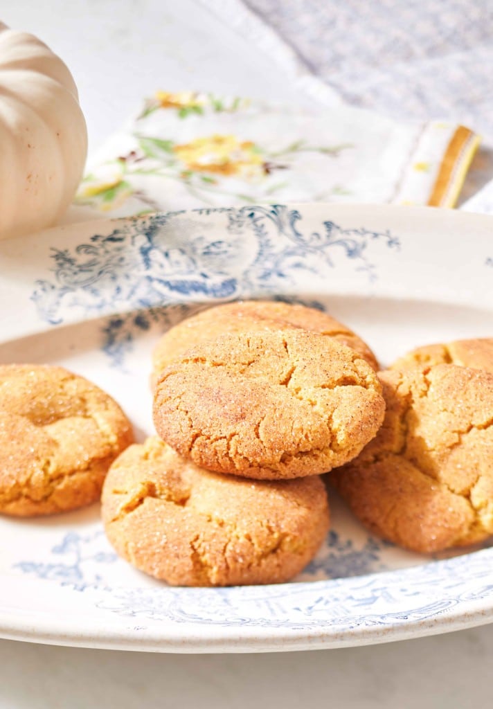 A decorative blue and white plate holds four golden-brown autumn cookies with a cracked surface. In the background, there is a small white pumpkin and a floral-patterned cloth napkin. The scene is bright and evokes a cozy, homemade feel.