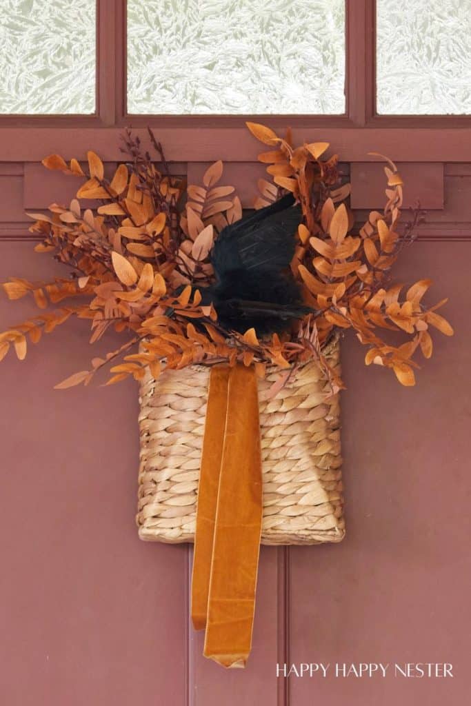 A woven basket with handles is filled with brown and orange foliage and a black decorative element. The basket, adorned with an orange ribbon, showcases festive fall decor and is hung on a brown door. Text at the bottom right reads "HAPPY HAPPY NESTER.