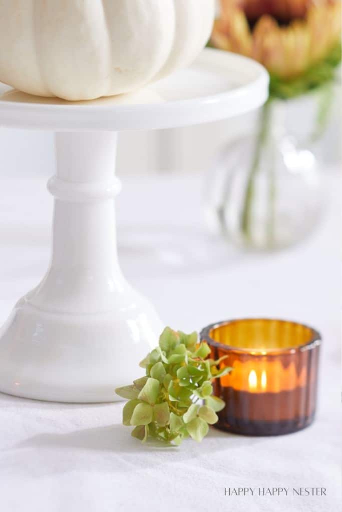 A white cake stand with a white pumpkin sits on a light-colored surface. Below it, a small green hydrangea flower rests next to an amber glass candle holder with a lit candle inside. A vase with blurred flowers is in the background.