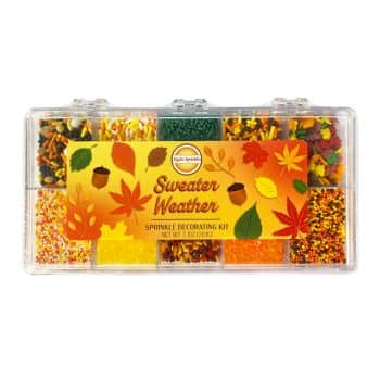 A clear plastic container labeled "Sweater Weather Sprinkle Decorating Kit" displays various compartments filled with colorful, autumn-themed sprinkles in shades of yellow, orange, red, green, and brown. Perfect for decorating autumn cookies, this kit features shapes like leaves and acorns.