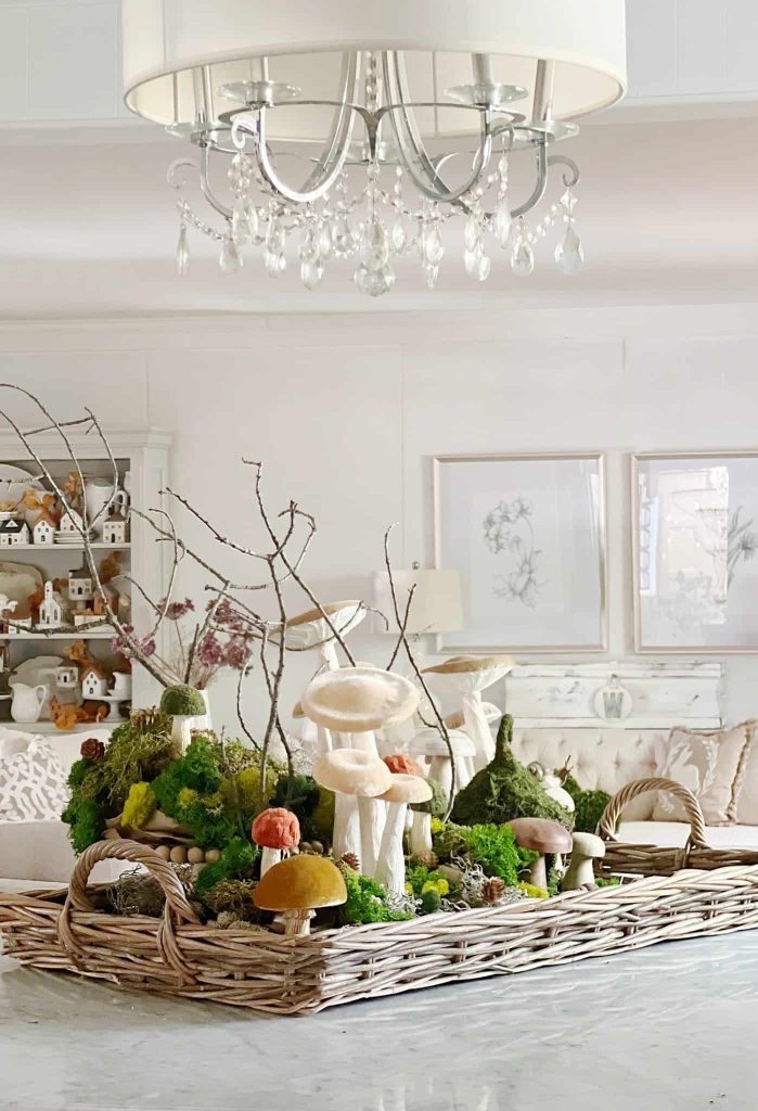 A wicker basket centerpiece on a white table holds an arrangement of faux mushrooms, green moss, and twigs. Perfect for fall decor ideas, the cozy living room in the background features white walls, framed botanical prints, and small house decorations on shelves. A chandelier hangs above.