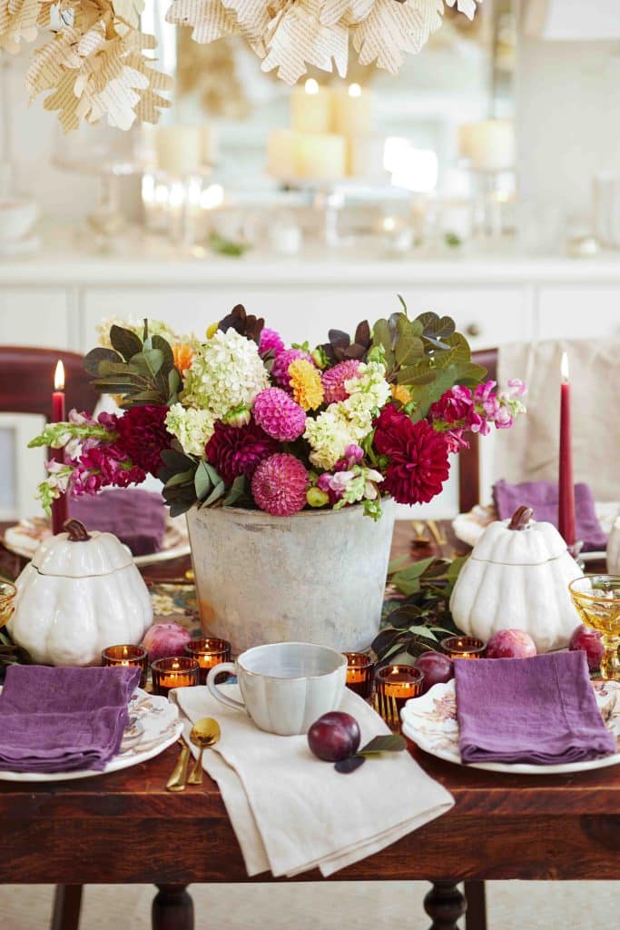 A fall-themed dining table features purple napkins, gold flatware, white plates, and ceramic pumpkin decorations. For those seeking fall decor ideas, the centerpiece boasts a metal pot filled with a colorful assortment of flowers. Two tall candles and small votives add a warm ambiance.