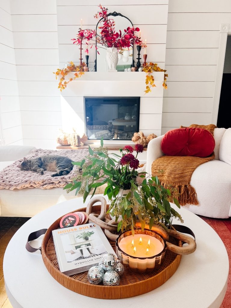 A cozy living room with a white fireplace adorned with festive fall decor, including red and orange leaves. A cat is sleeping on a knitted blanket by the fire. A round coffee table features a lit candle, a vase with flowers, a magazine, and Christmas ornaments.