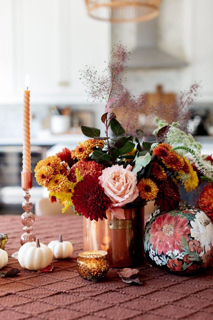 A copper vase filled with a large, colorful arrangement of roses, chrysanthemums, and various autumn flowers serves as the centerpiece on a textured brown tablecloth. Surrounding it are small white pumpkins, a floral-patterned pumpkin, a lit taper candle, and a gold textured votive candle.