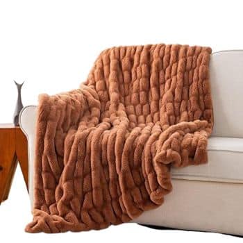 A fluffy, quilted brown blanket draped over the arm and back of a white sofa. The blanket features a thick plush texture. A wooden side table with a sleek, gray decorative item is visible beside the sofa.