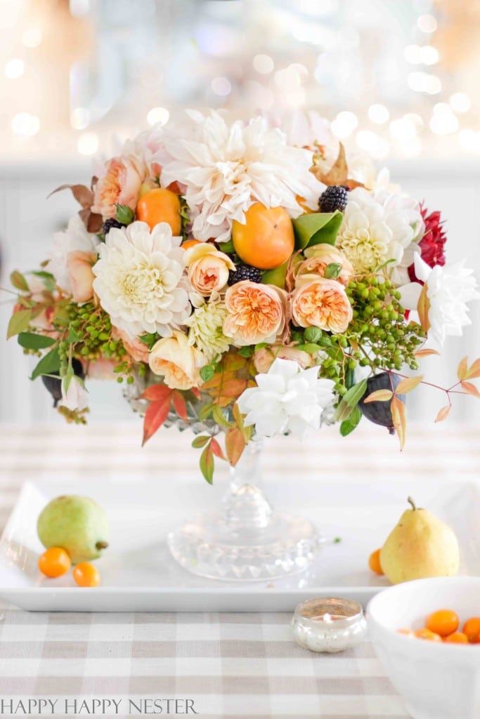 A floral arrangement with peach and white flowers, greenery, and small orange fruits in a clear glass vase on a checkered tablecloth. Surrounding the vase are a pear, a small bowl of kumquats, and a tealight candle. Perfect for fall decor ideas, the background is softly blurred with lights.