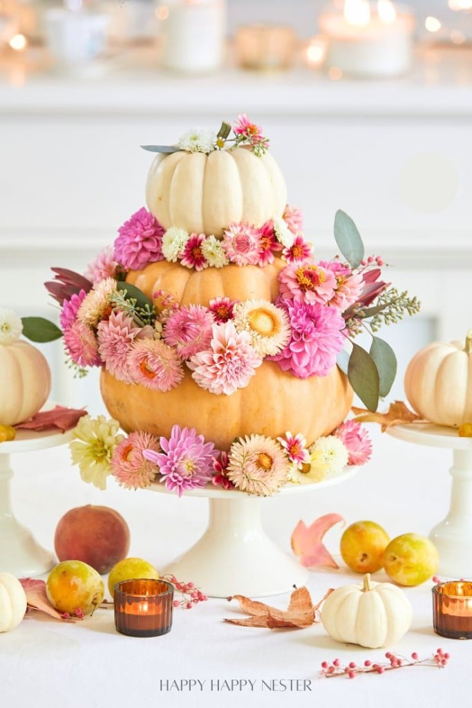 Create your own DIY autumn centerpiece with a three-tiered arrangement of white and orange pumpkins adorned with pink, white, and peach flowers and green leaves, set on a white cake stand. Surrounding the pumpkin centerpiece are small pumpkins, leaves, and lit candles.