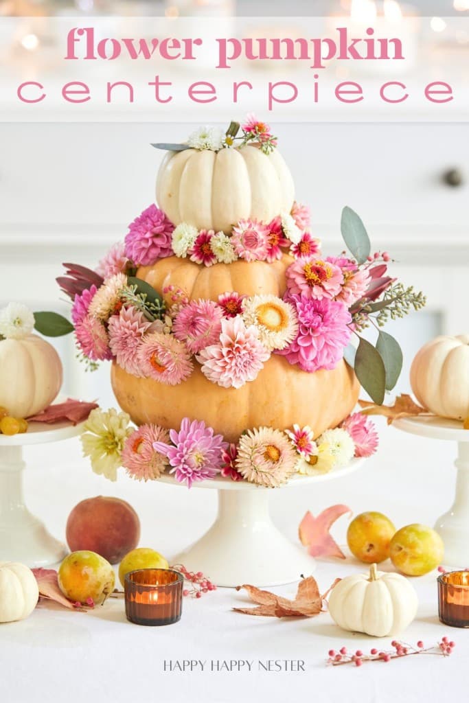 A tiered pumpkin centerpiece decorated with pink, white, and yellow flowers is displayed on a white table. Smaller pumpkins, candles, and autumn leaves surround it. The text above reads "flower pumpkin centerpiece.