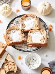 Slices of raisin bread with ghost-shaped icing lay on a decorative plate surrounded by small pumpkins, candles, candy corn, and a bowl of candy eyes. A ghost-shaped cookie cutter and a wooden cutting board are also visible.