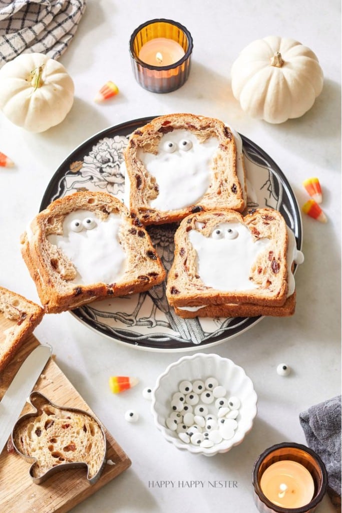 A plate filled with slices of toast that have ghost shapes made of icing, complete with candy eyes, is surrounded by small white pumpkins, candy corn, candles, and scattered edible eyes on a marble surface. Halloween-themed baking and autumn recipes are displayed on a festive plate.