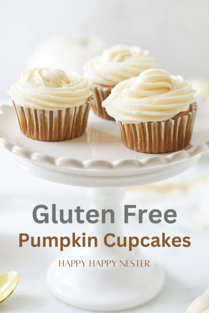 A decorative white cake stand displays three gluten-free pumpkin cupcakes topped with smooth swirls of white frosting. The image text reads "Gluten Free Pumpkin Cupcakes" with "Happy Happy Nester" below. The background is softly blurred, highlighting the cupcakes.