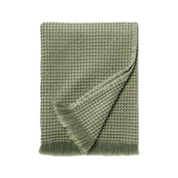 A neatly folded, light green waffle-knit blanket with fringed edges is displayed against a plain white background. The textured surface and draped fringe give it a cozy and stylish appearance.