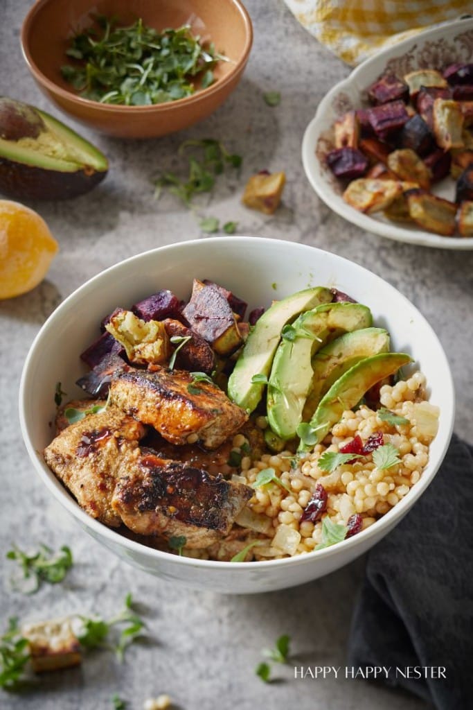 A white bowl filled with grilled meat, avocado slices, roasted vegetables, and couscous, garnished with herbs. Surrounding it are a bowl of mixed greens, a sliced avocado, and a plate of roasted vegetables—perfect for autumn recipes.