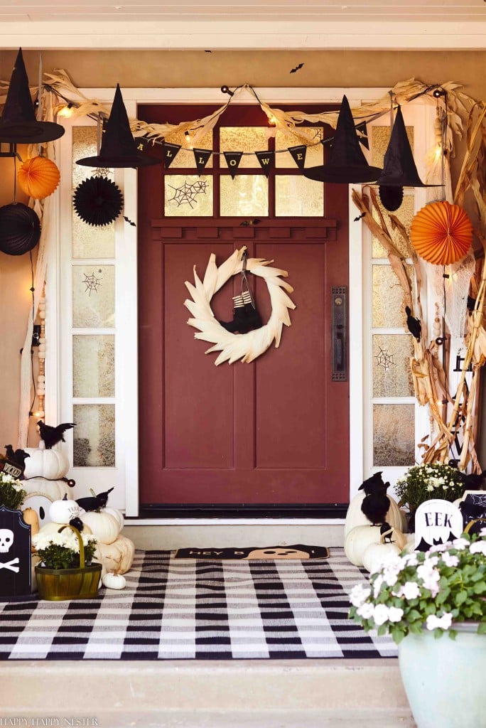 A front porch decorated for Halloween features a burgundy door with a white wreath, black witch hats hanging above, and a "HALLOWEEN" banner. White pumpkins and spooky decorations are arranged around the entrance. For fall decor ideas, a black and white checkered rug lies on the floor.