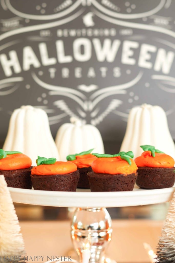 A dessert display features mini cakes with orange frosting resembling pumpkins on a glass cake stand. Black and gold Halloween decor accents the scene, complemented by decorative white pumpkin ornaments and a festive "Halloween Treats" sign in the background.