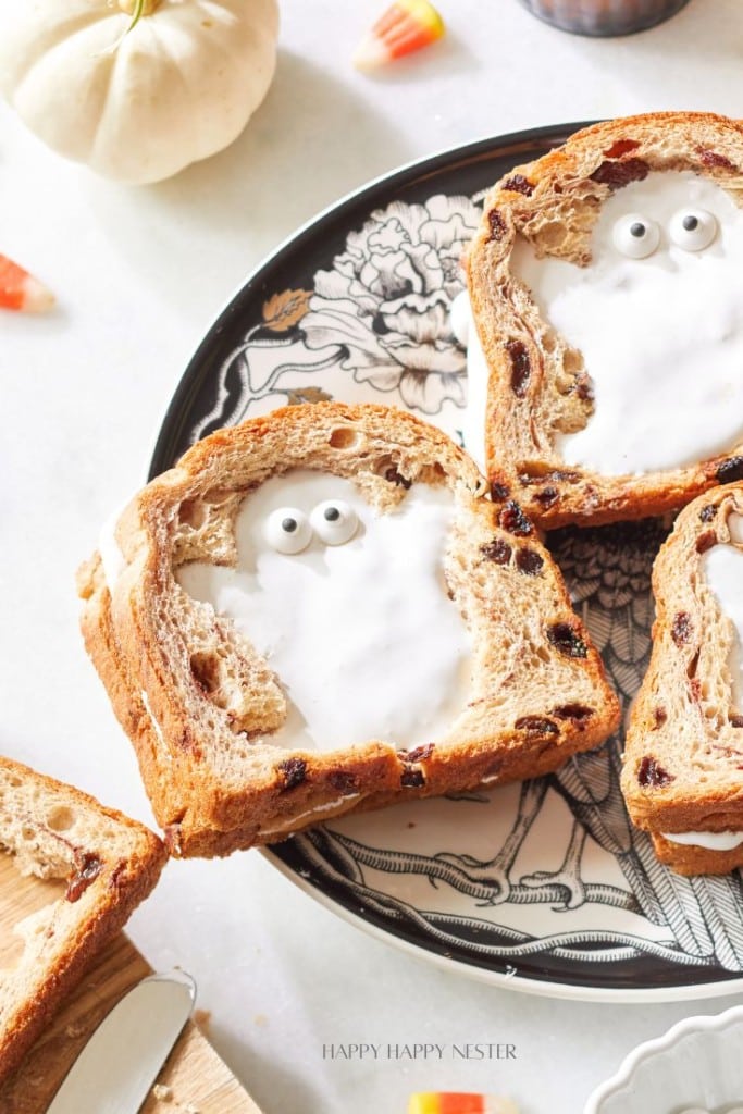 Two slices of raisin bread with white frosting and googly eyes arranged to resemble ghosts are placed on a Halloween-themed plate. A small white pumpkin and candy corn pieces are visible in the background.