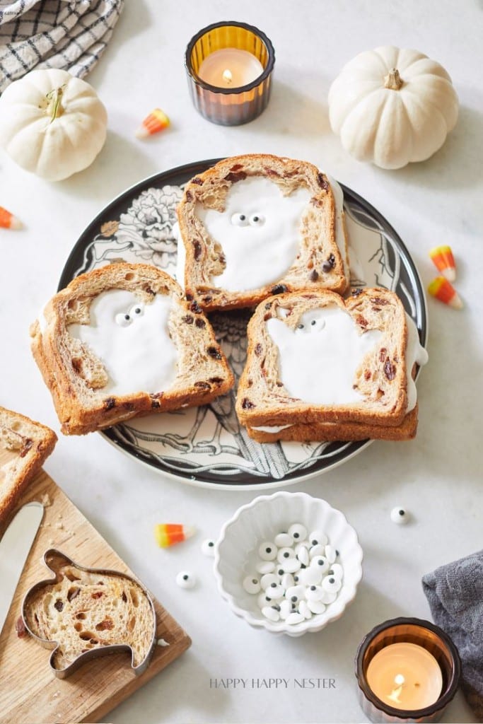 Three slices of toast feature ghost shapes made of icing on a decorative plate, surrounded by small pumpkins, candy corn, googly eyes, and lit candles. A ghost-shaped cookie cutter, additional toast slices, and a gray cloth napkin are also visible.