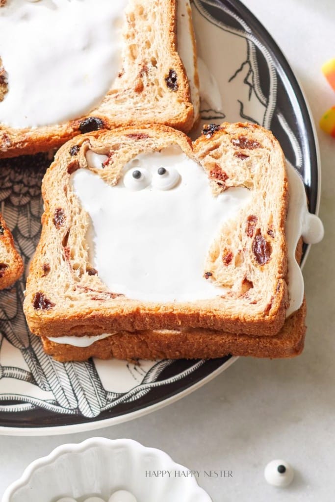 Two slices of raisin bread stacked with a ghost-shaped frosting in the center, topped with candy eyes on a decorative black and white plate. In the background, other partially visible bread slices and candy eyes are present.