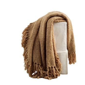 A cozy, light brown textured blanket with fringe edges is draped over a white cushion or chair. The blanket's fabric appears soft and inviting.