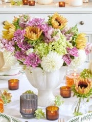 A beautifully set Thanksgiving table featuring a white vase filled with a colorful flower arrangement of pink, yellow, and white blooms. Surrounding the centerpiece are small pumpkins, candles in glass holders, and elegant plates and cutlery.