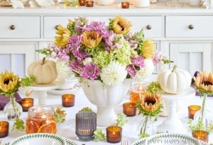 A beautifully set Thanksgiving table featuring a white vase filled with a colorful flower arrangement of pink, yellow, and white blooms. Surrounding the centerpiece are small pumpkins, candles in glass holders, and elegant plates and cutlery.