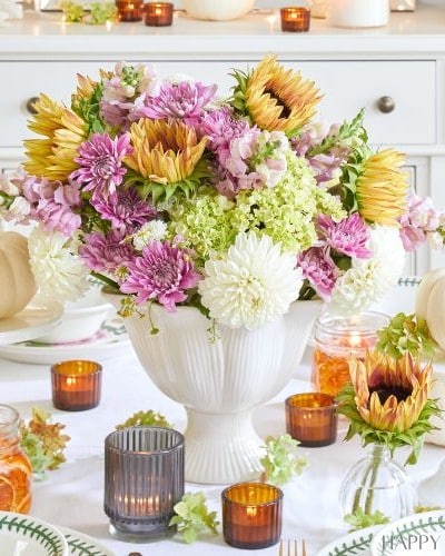 A beautifully set Thanksgiving table featuring a white vase filled with a colorful flower arrangement of pink, yellow, and white blooms. Surrounding the centerpiece are small pumpkins, candles in glass holders, and elegant plates and cutlery.