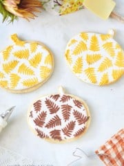 Three pumpkin-shaped cookies are decorated with intricate fern patterns in yellow, gold, and brown icing. The cookies are placed on a white marble surface, surrounded by autumn-themed decor, including a sunflower, a spatula, and a checked cloth.