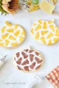 Three pumpkin-shaped cookies are decorated with intricate fern patterns in yellow, gold, and brown icing. The cookies are placed on a white marble surface, surrounded by autumn-themed decor, including a sunflower, a spatula, and a checked cloth.