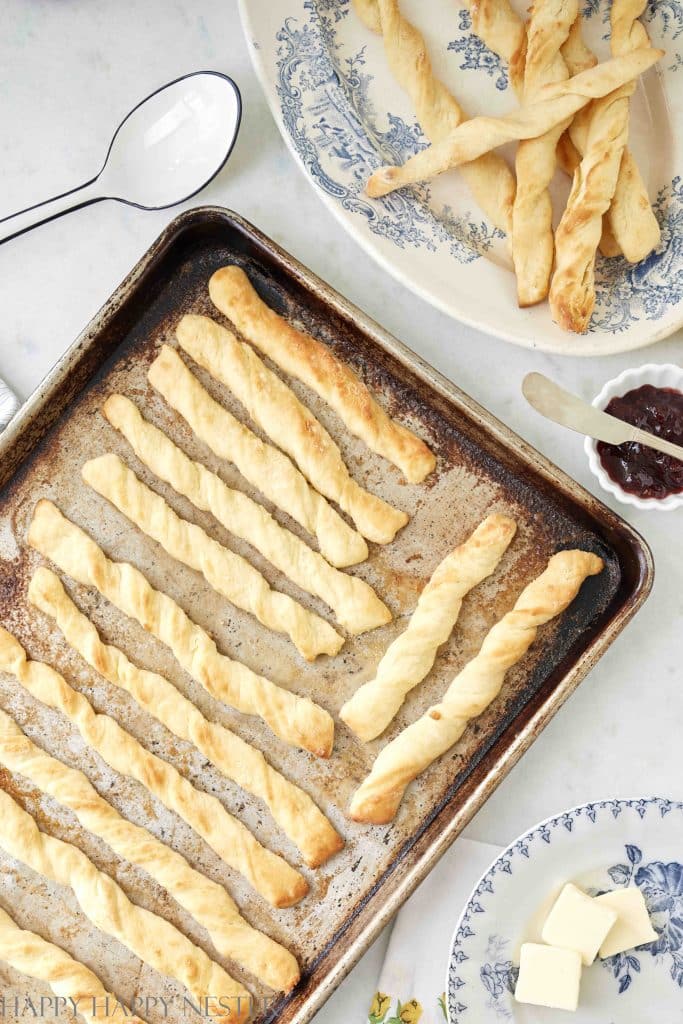 A baking tray filled with baked twisted breadsticks sits on a marble countertop. Next to the tray is a plate with more breadsticks and a spoon with dark jam. Also visible are a plate with butter cubes and a decorative plate, perfect for autumn recipes in a light and airy kitchen setting.