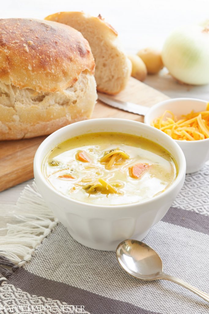 A white bowl filled with creamy soup containing vegetables like carrots and broccoli sits on a grey tablecloth alongside a metal spoon. Behind the bowl, there is a loaf of bread, a slice of cheese, an onion, and a potato on a wooden cutting board—perfect for cozy autumn recipes.