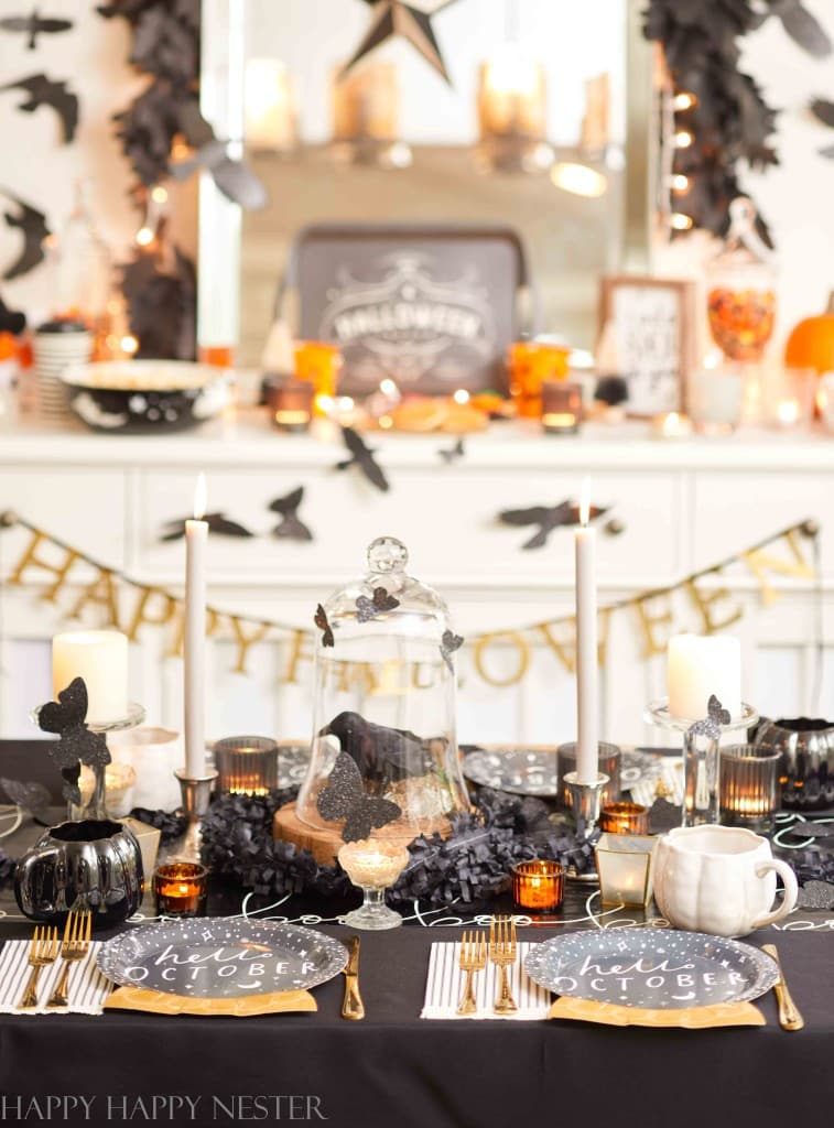 A spooky Halloween-themed table arrangement featuring black and white decorations is perfect for those looking for fall decor ideas. Plates with "hello OCTOBER" on them, candles, and black painted pumpkins create a festive backdrop. A "HAPPY HALLOWEEN" banner and eerie details like crows and cobwebs add to the ambiance.