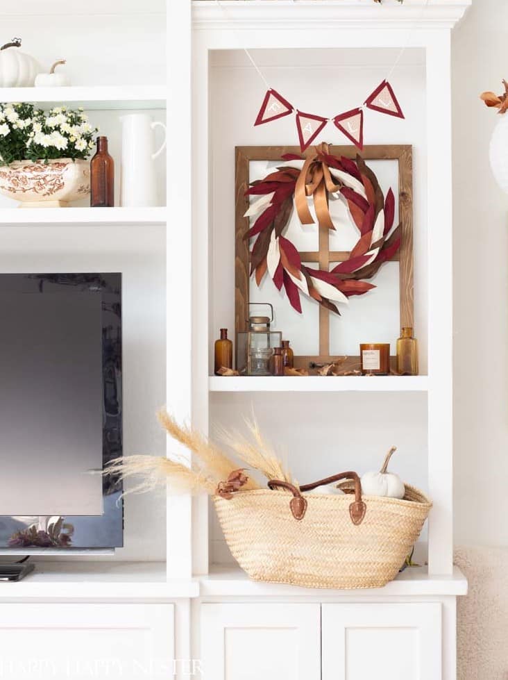 A decorated shelf with festive fall decor: a straw basket containing dried wheat and pumpkins at the bottom, a TV to the left, a mirror with a fall wreath and amber bottles in the center, and a white jug with flowers and additional decorations on the top shelf. A triangular pennant banner hangs above.