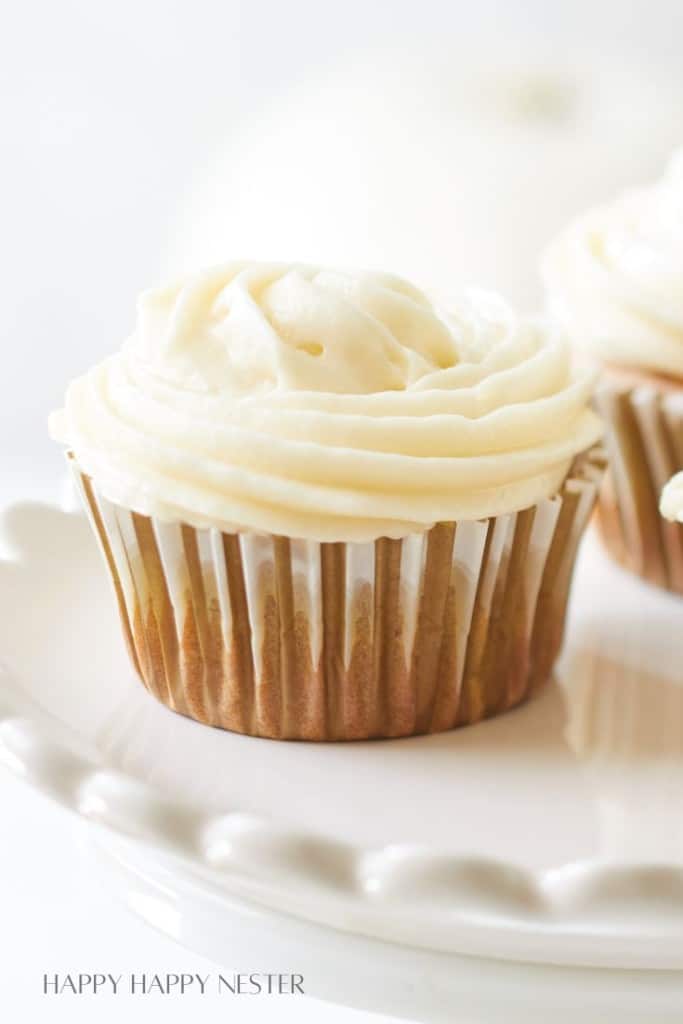 A close-up of a single cupcake with white frosting swirled on top, displayed on a white stand. The cupcake liner is striped in shades of brown and white. The background is blurred, highlighting the cupcake as the main focus.