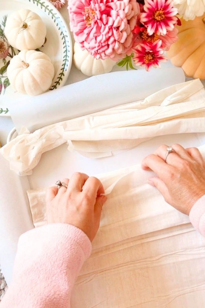 Close-up of hands in a pink sweater folding light-colored dough strips on a parchment-lined surface. The background includes a plate with small white pumpkins, fresh pink and red flowers, and other light fall decor.