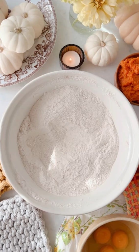 A large white mixing bowl filled with flour is surrounded by ingredients for baking, including a bowl of eggs, a bowl of pumpkin puree, and a lit candle. White pumpkins and flowers are arranged on a decorative plate nearby.