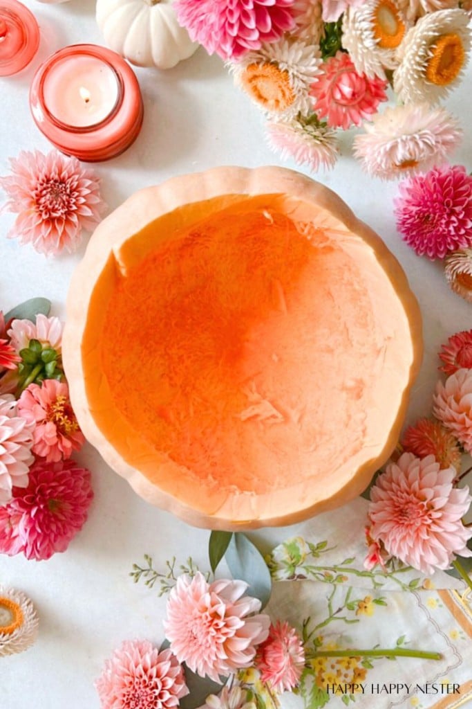 A carved-out pumpkin surrounded by pink and white flowers, with two pink candles in glass holders placed nearby. The pumpkin is hollow and has a smooth, orange interior. The scene is bright and colorful, giving a festive and cozy autumn atmosphere.