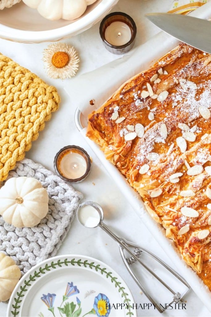 A cozy scene featuring a freshly baked almond pastry on a tray, topped with sliced almonds and powdered sugar. Surrounding the tray are small white pumpkins, knitting, candles, a floral plate, and a spoon, creating a warm, inviting atmosphere.