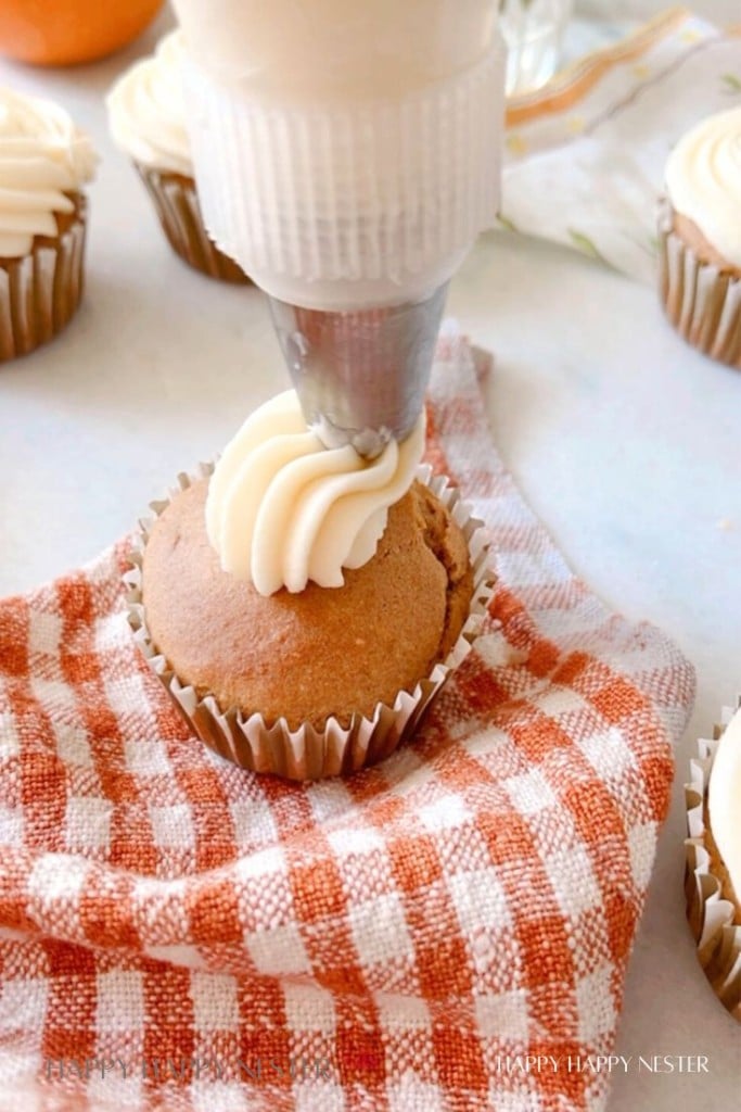 A close-up image of a cupcake being decorated with white frosting using a piping bag. The cupcake is placed on an orange and white checkered cloth, and other undecorated cupcakes can be seen in the background.