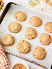 A baking sheet with a parchment paper lining holds nine freshly baked peanut butter cookies arranged in rows. In the lower left corner, a white bowl of brown sugar and a checkered cloth are visible. The scene is bright and has an autumnal feel with leafy decor.