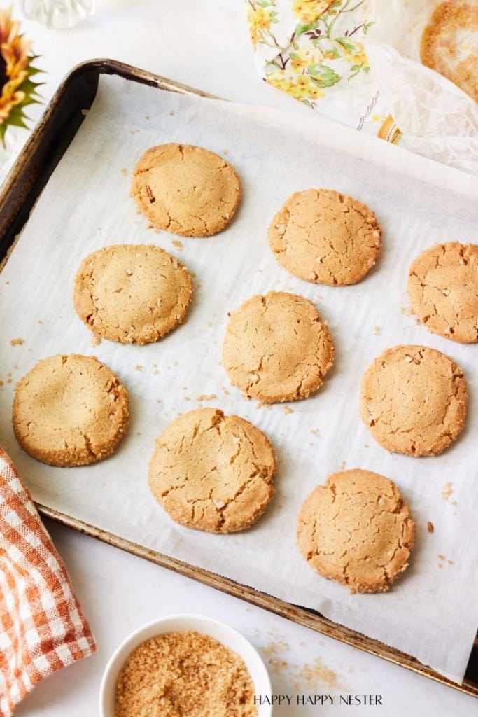 A baking sheet with a parchment paper lining holds nine freshly baked peanut butter cookies arranged in rows. In the lower left corner, a white bowl of brown sugar and a checkered cloth are visible. The scene is bright and has an autumnal feel with leafy decor.