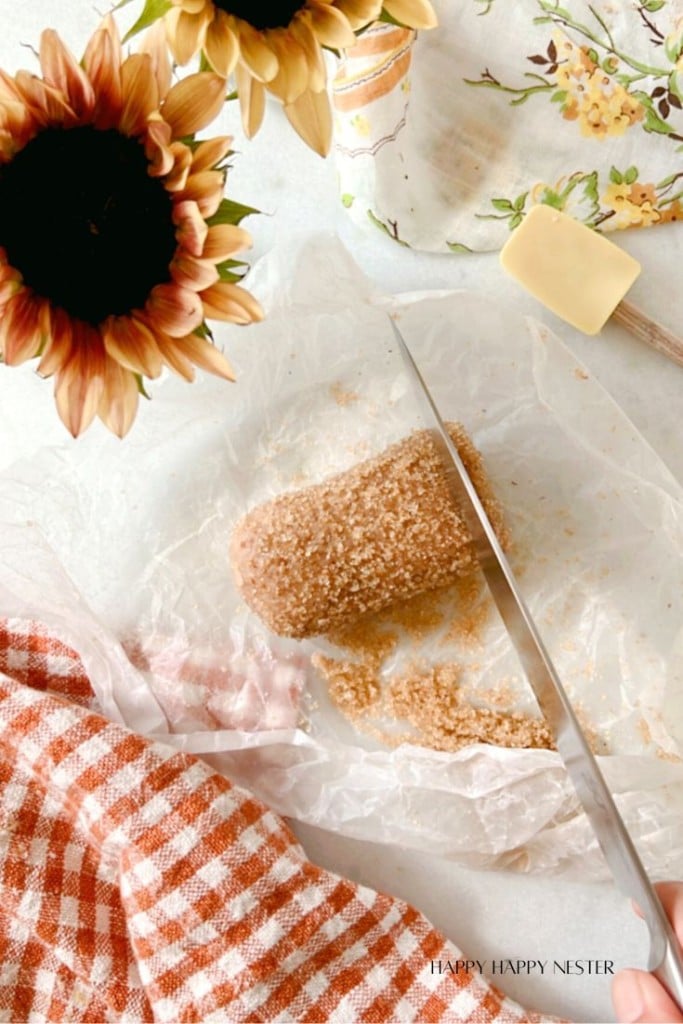 A block of brown sugar on parchment paper is being sliced with a knife. Sunflowers and a floral-patterned cloth are in the background, along with a yellow spreading knife. A checkered orange and white cloth is partially visible in the foreground.