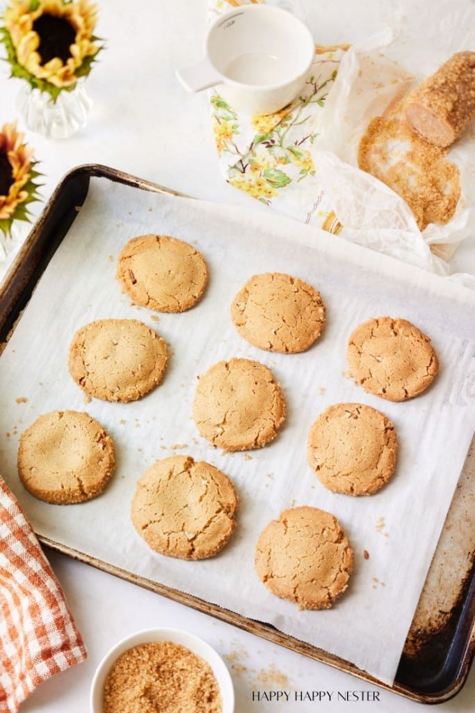 A baking tray with nine freshly baked cookies on a sheet of parchment paper. The tray sits on a white surface accompanied by a red-checkered towel, a bowl of brown sugar, a floral cup, a roll of parchment paper, and a vase of sunflowers.